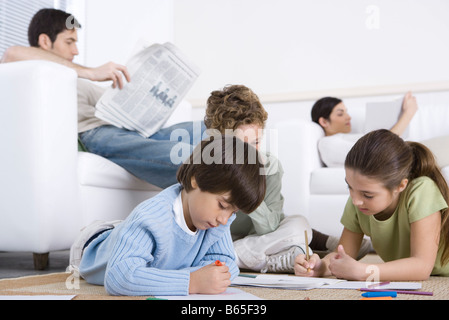 Children coloring on the floor in living room, parents reading in background Stock Photo
