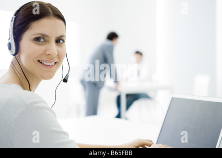 Woman wearing headset, working at desk, smiling over shoulder at camera Stock Photo