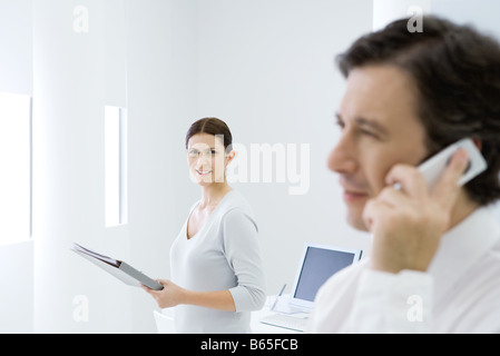 Professional woman in office, smiling at camera, male colleague using cell phone in foreground Stock Photo