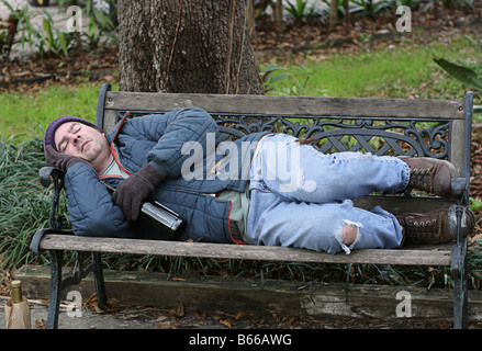 A full view of a homeless man asleep on a park bench with his wine bottle Stock Photo