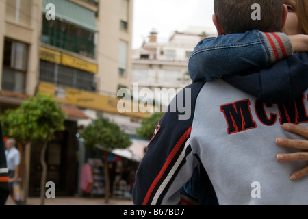 A couple holding and kissing next to a building detailed male jacket with red and white strips. The girl is kissing him warmly Stock Photo