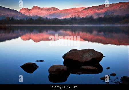 Dawn light illuminates the Langdale Pikes with a rosy glow reflected in the still waters of Elterwater Lake District England UK