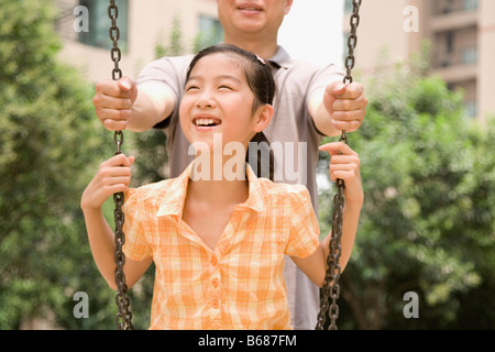 Girl smiling on a swing with her father standing behind her Stock Photo