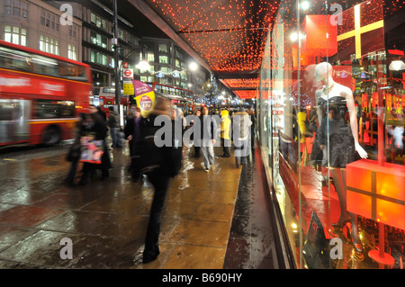 Shoppers on wet pavement outside department store front window Xmas gifts on display Oxford Street evening Christmas lights rain in London West End UK Stock Photo