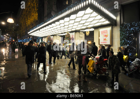 Night view busy Oxford street shoppers & tourists silhouette & Christmas shopping in the rain outside Selfridges department store entrance London UK Stock Photo