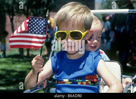 A young boy wearing sun glasses and his little brother ride in a carriage and wave an American flag at a 4th of July Parade. Stock Photo