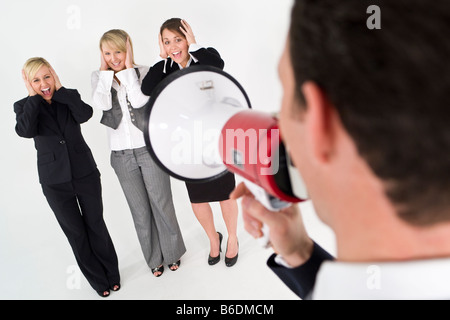 A businessman shouts through a megaphone while three business woman cover their ears and scream Stock Photo