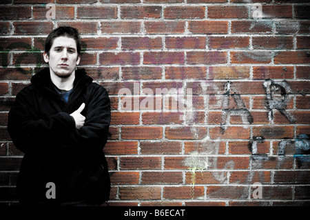 Young man leaning on a graffiti covered brick wall, arms crossed, looking aggressive UK Stock Photo