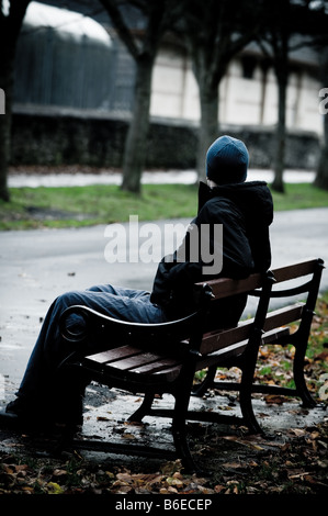 Young man wearing a hooded jacket alone and isolated sitting on a park bench Stock Photo