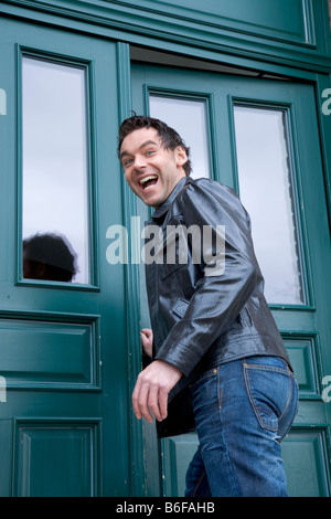 Young man laughing, wearing a leather jacket and entering a green door Stock Photo