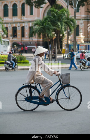 A man wearing traditional Vietnamese clothes and conical hat, cycling along a street in central Ho Chi Minh City, Vietnam Stock Photo