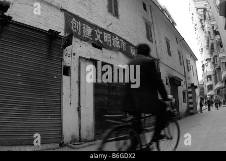 Chinese man riding bike through urban side streets of old Canton past old warehouse in residential neighborhood under propaganda Stock Photo