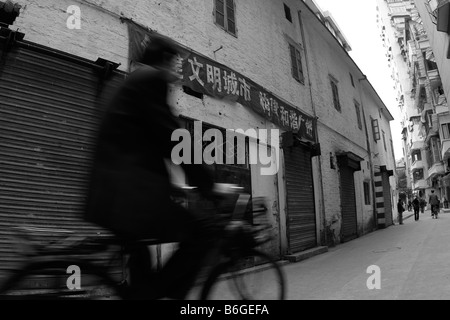 Chinese man riding bike through urban side streets of old Canton past old warehouse in residential neighborhood under propaganda Stock Photo