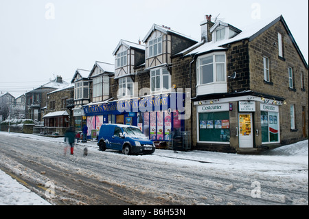 The local village shops on high street after a fall of snow - people walking on snowy pavement & road - Burley in Wharfedale, Yorkshire, England, UK. Stock Photo