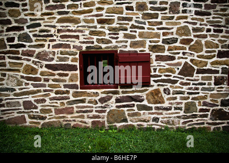 Ventilation window, painted red, on the side of an old Colonial American stone barn surrounded by green grass. Stock Photo