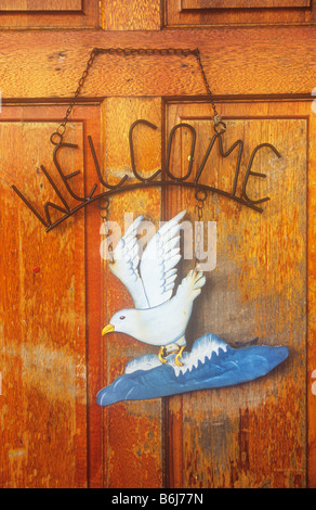 Metal sign hanging by chain on weathered varnished door stating Welcome and with cutout tin image of painted seagull on waves Stock Photo