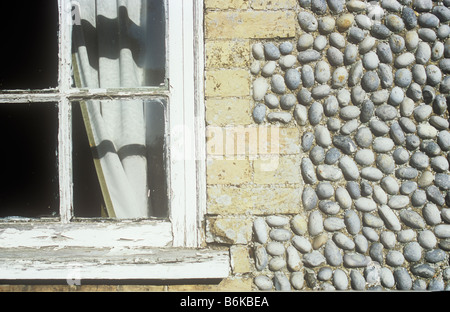 Detail of window frame with curtain and shadow and cracked white paint with pale brick surround set in neat flint wall Stock Photo