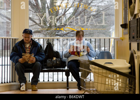 Elderly couple sit together and read as they wait for their laundry at launderette, laundromat. Stock Photo