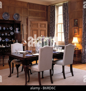 Upholstered beige highback chairs and antique table in panelled country dining room with lighted lamp on small side table Stock Photo