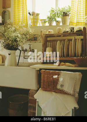 Plates on wooden rack above basket of old linen in cottage kitchen with yellow checked curtains above white jug in white sink Stock Photo