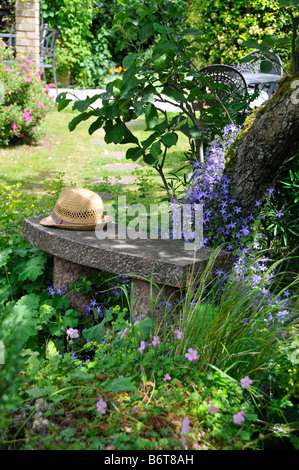 Traditional English garden scene with straw hat on bench under tree surrounded by flowers Stock Photo