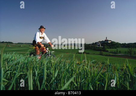 Man wearing traditional costume riding a bicycle, Andechs Abbey in background, Bavaria, Germany Stock Photo