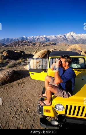 Man sitting on a jeep in the desert next to mountains with binoculars. Stock Photo