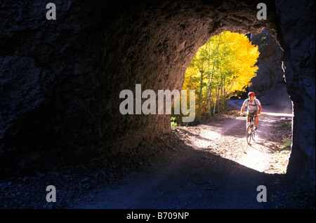 A young man pedals his mountain bike past golden aspen leaves and into a rock tunnel while mountain biking in the San Francisco Stock Photo