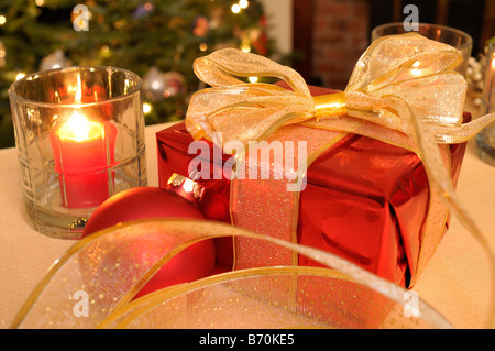 Gift wrapped red Christmas present with baubles lighted tree candles and gold ribbon on table. Stock Photo