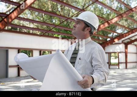 A construction inspector holding blueprints and looking at the roof beams of a steel building in progress Stock Photo