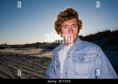 Young man wearing denim jacket standing on beach at sunset Stock Photo