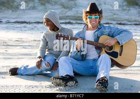 Young interracial couple sitting at beach, man playing guitar Stock Photo