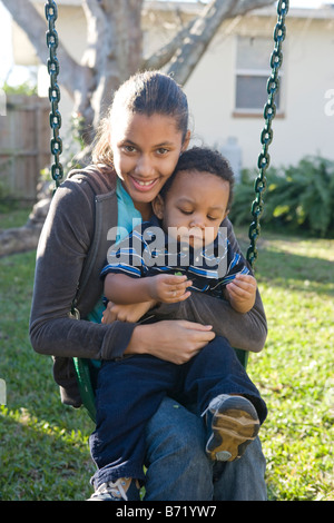 African American pre-teen girl sitting on swing holding baby brother in backyard Stock Photo