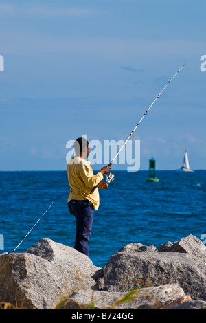 Palm Beach Shores , African American middle aged man fishing from jetty  on rocks with sailing yacht and buoy in deep blue sea Stock Photo