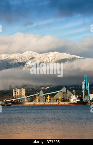 Cargo ship in Vancouver harbour in front of snow covered mountains with white clouds and a blue sky. Stock Photo