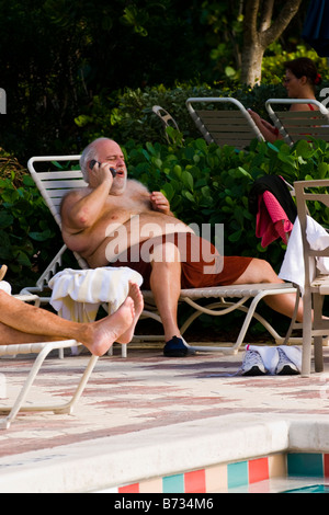 Palm Beach Shores , swimming pool , gray haired large middle aged man on lounger in swimming shorts talks on mobile cell phone Stock Photo