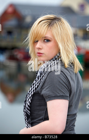 blonde 18 year old girl looking over her shoulder towards camera wearing t shirt and scarf in front of carrick harbour Stock Photo