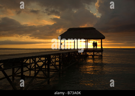 Man and woman standing on the end of a wooden pier holding hands at sunset over the Indian Ocean Mauritius Stock Photo