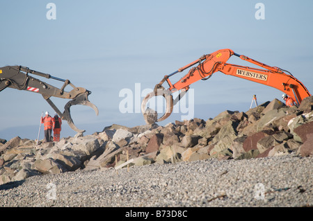Mechanical JCB diggers placing large boulders repairing the sea defences on the beach at Tywyn Gwynedd north wales UK Stock Photo