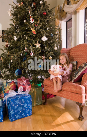 Young girl sitting in chair next to Christmas tree and presents Stock Photo