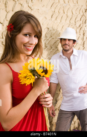 Young woman holding flower while young man watches in background Stock Photo