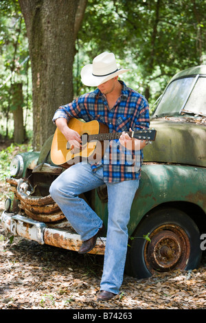 Young man wearing cowboy hat playing guitar leaning on old car Stock Photo