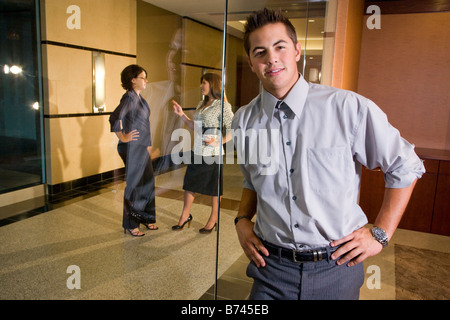 Office worker leaning on glass wall, with co-workers chatting outside in corridor Stock Photo
