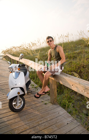 Young man sitting on railing of wooden boardwalk by parked moped Stock Photo