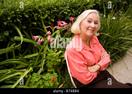 Mature woman sitting on chair on sidewalk in front of flowers and hedges Stock Photo