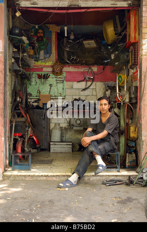 A young mechanic sits in his Shahr-e Rey bike shop in Tehran, Iran, in January 2009, with an Imam Ali portrait poster on the back wall. Stock Photo