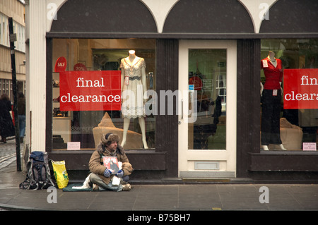 Homeless man trying to sell Big Issue magazines outside a clothes shop in cold weather and snow. Stock Photo