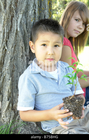 13 year old girl and six year old boy with small tree seated beside tree, Winnipeg, Canada Stock Photo