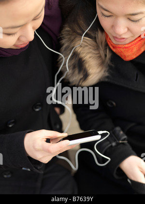 Two young woman share headphones while listening to music on a smart phone. Stock Photo