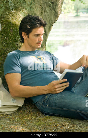 Man sat down leaning against a tree reading a book Stock Photo